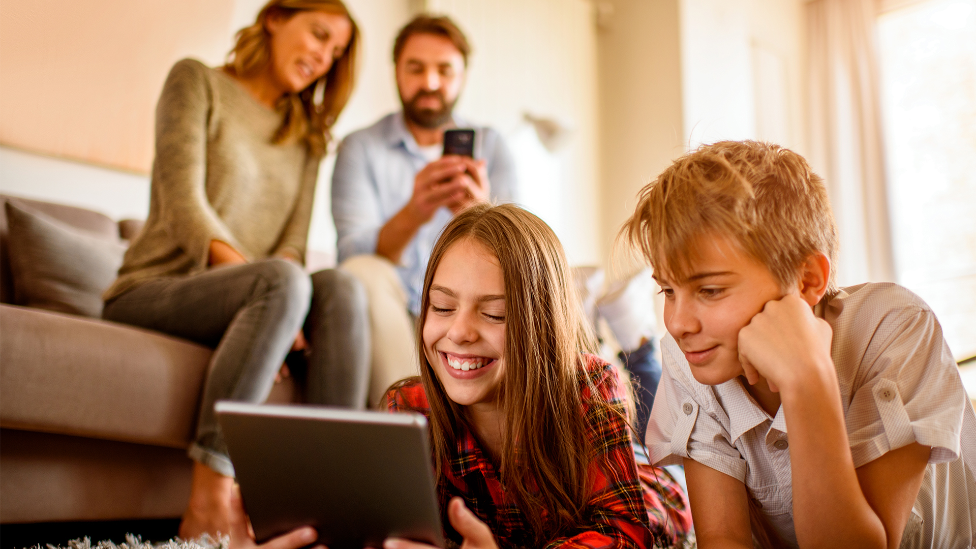 Brother and sister using digital tablet on floor while their parents sitting on sofa behind them.