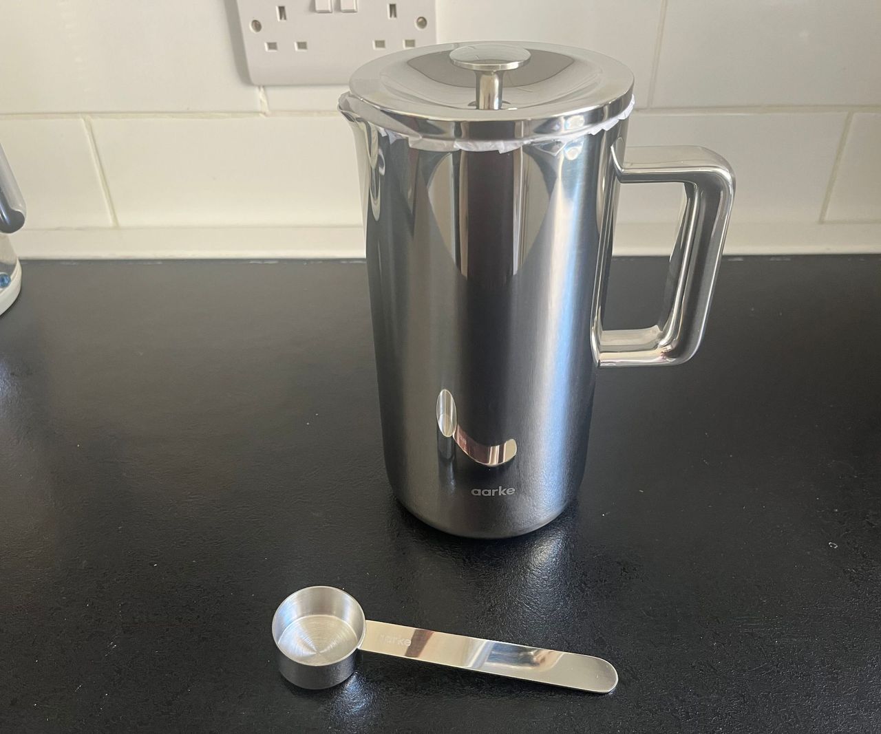 A stainless steel coffee press on a black counter, with a stainless steel coffee scoop