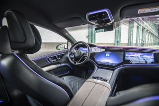 A photo of the Mercedes Benz EQS interior overlooking the driver seat and dashboard. The interior is black with a leather seat. The dashboard feature a dashboard-wide display.