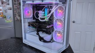 iBUYPOWER RDY Trace X R01 pre-built gaming PC on a dark countertop shows vibrant RGB lighting inside, illuminating fans and components. The background features a fridge and door.