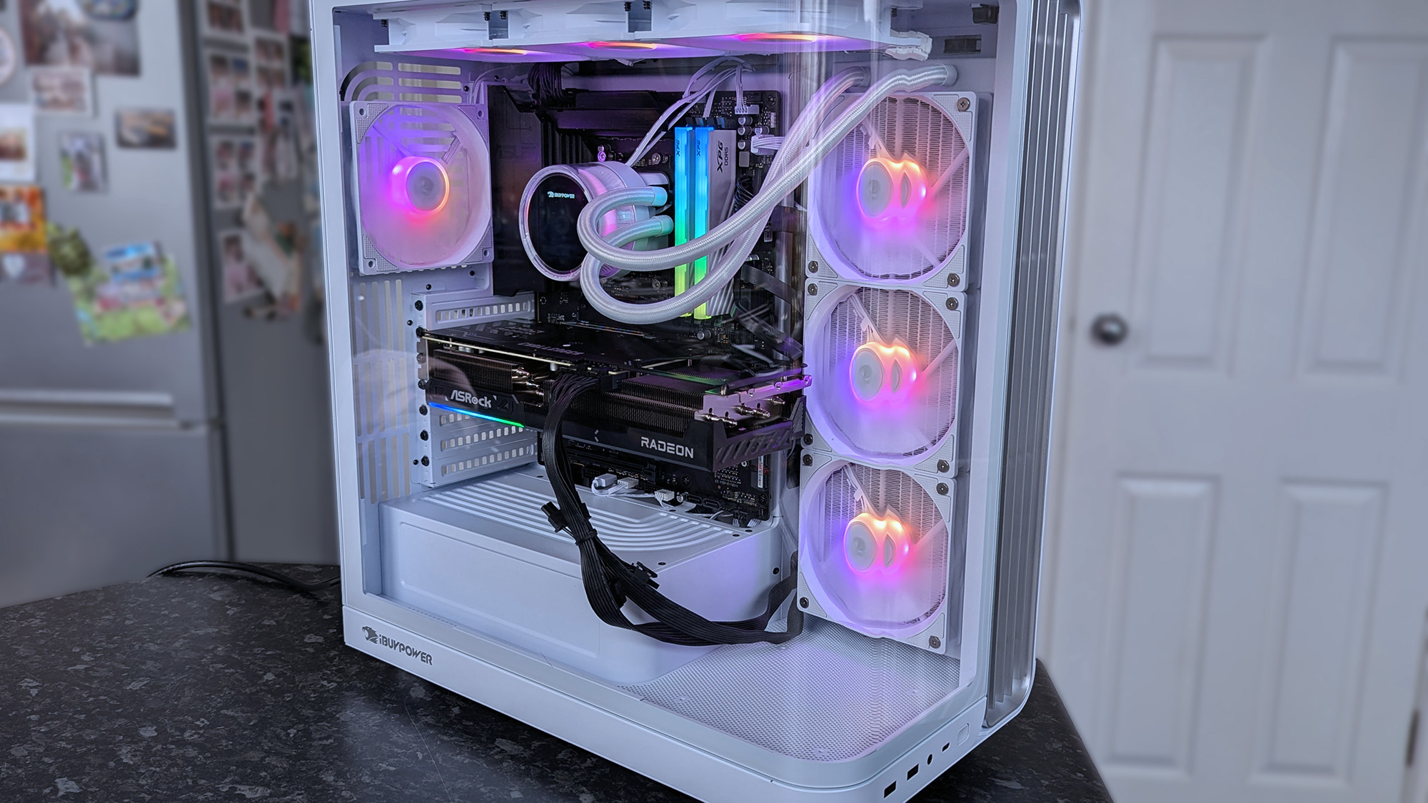 iBUYPOWER RDY Trace X R01 pre-built gaming PC on a dark countertop shows vibrant RGB lighting inside, illuminating fans and components. The background features a fridge and door.