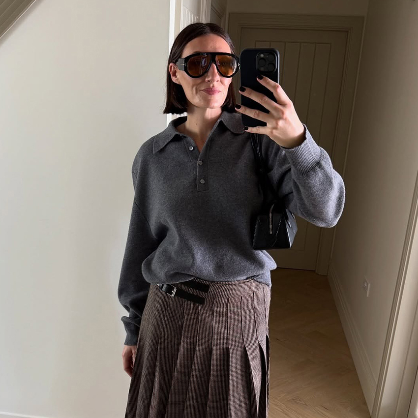 a woman wearing a gray sweater, a kilt plaid skirt, and round black sunglasses