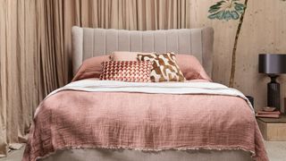 A bed with a pink linen throw in front of a wooden wall with a plant next to it