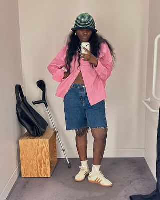 Woman wears long denim shorts, a knit bucket hat, pink button-down, and Adidas Samba sneakers.