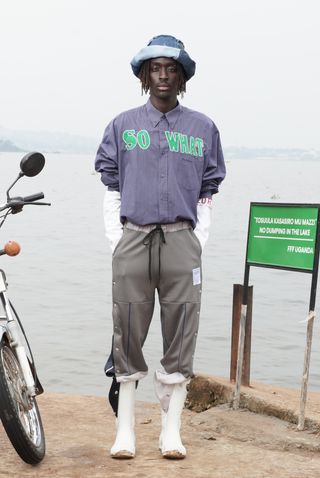 Man wearing 'So What' emblazoned shirt with popper. track pants and boots, standing next to body of water