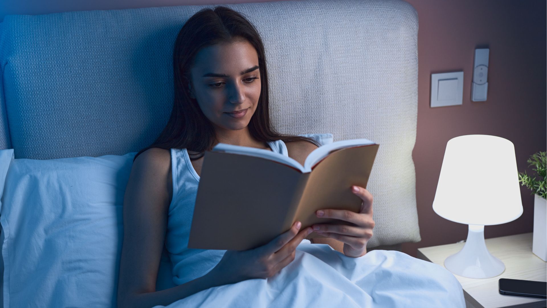 Woman reading in bed at night in a dimly lit room, with a lamp next to her providing light.
