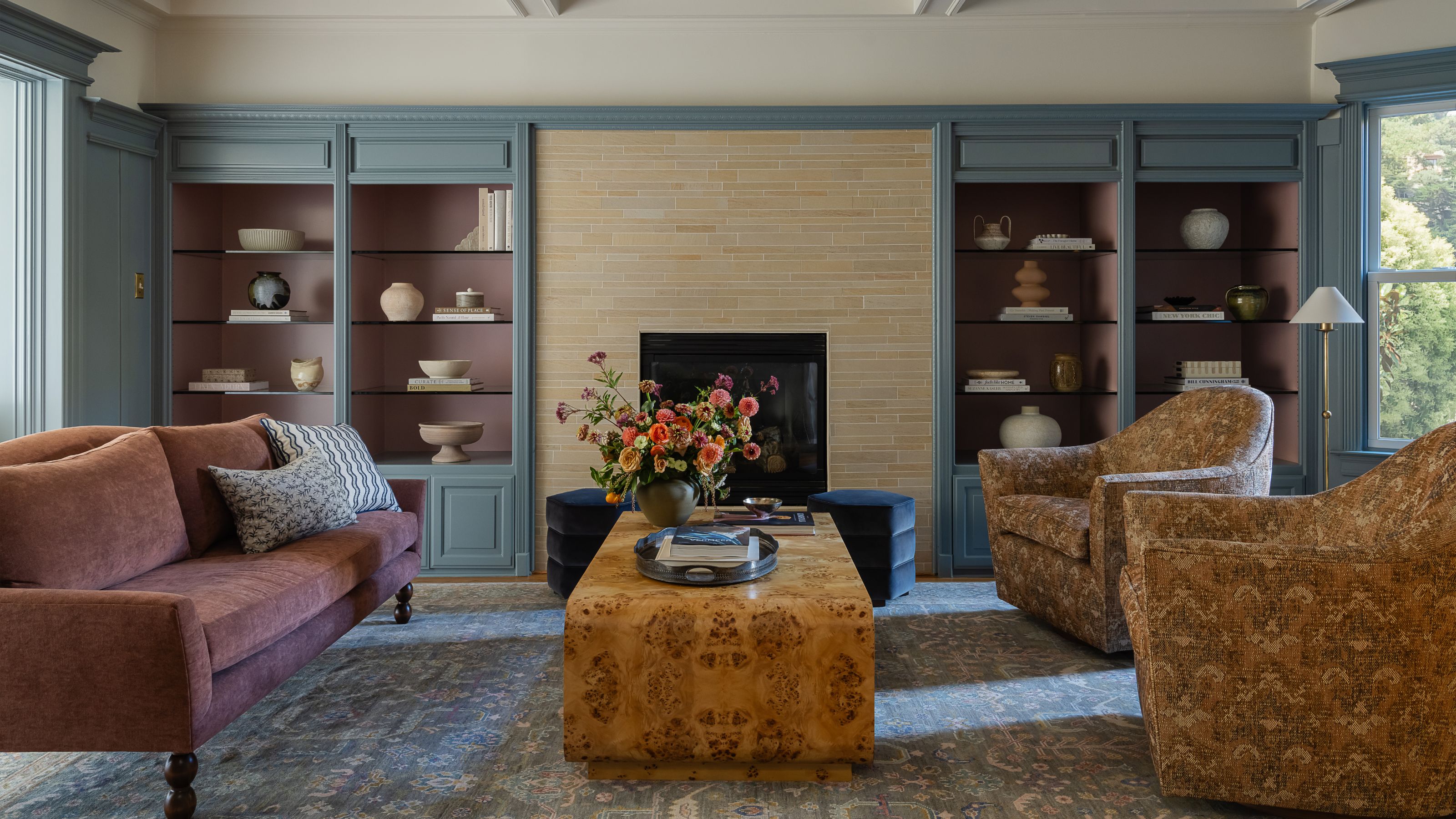 A sophisticated living room with a burl wood coffee table, a mauve sofa, and patterned armchairs, set against blue-grey built-in shelving flanking a fireplace