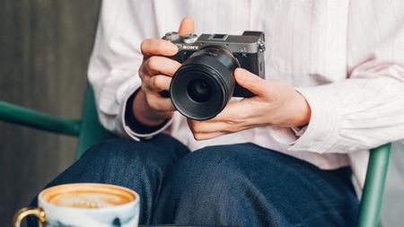 A person sitting in a green chair holds a silver and black Sony mirrorless camera with a prime lens over a cup of latte art.