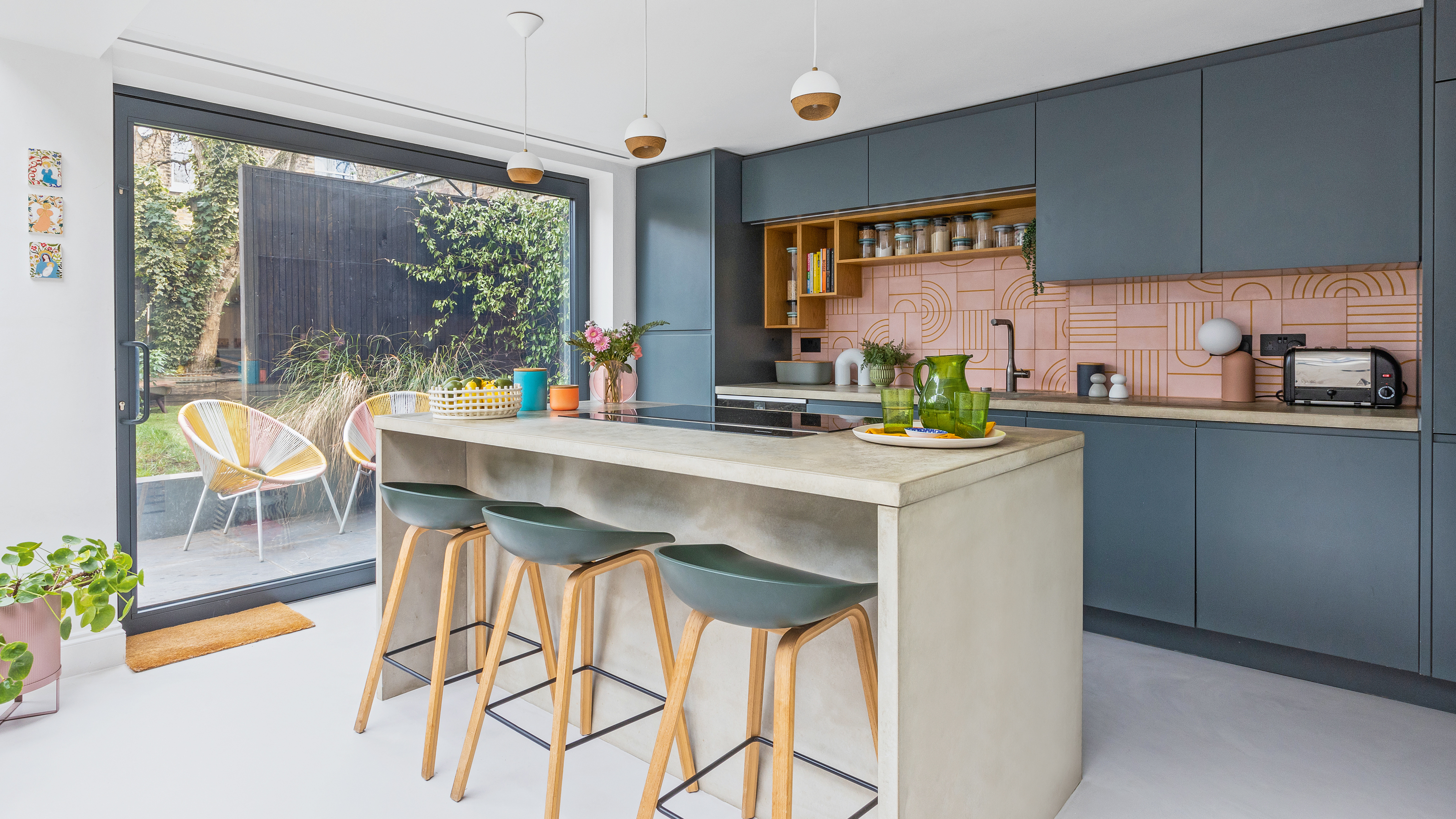 a contemporary dark blue kitchen with concrete kitchen island, bar stools and pendant lights with a pink graphic tiled splashback