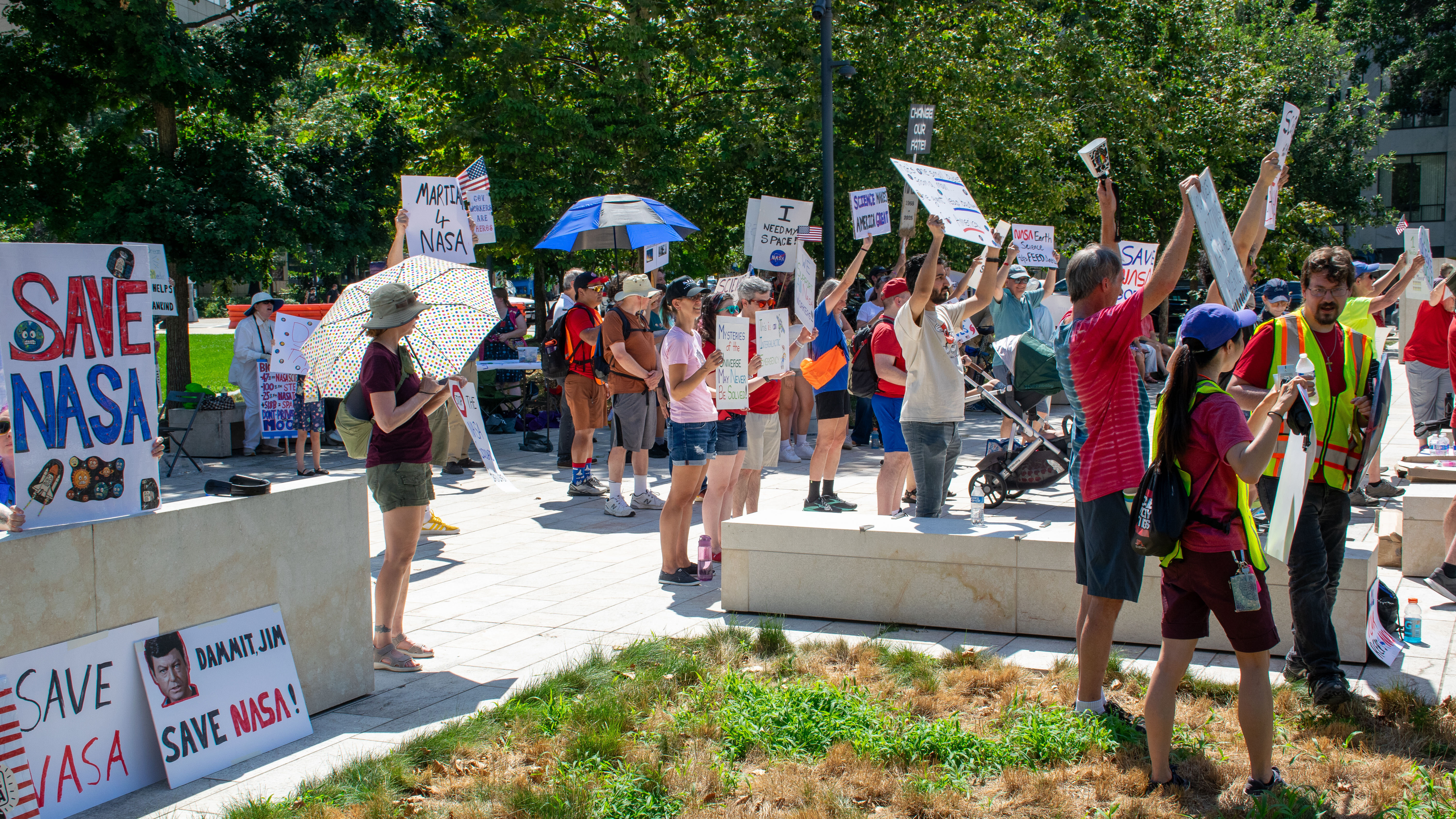 People hold signs outside on a sunny day in protest of NASA budget cuts, July 20, 2025.
