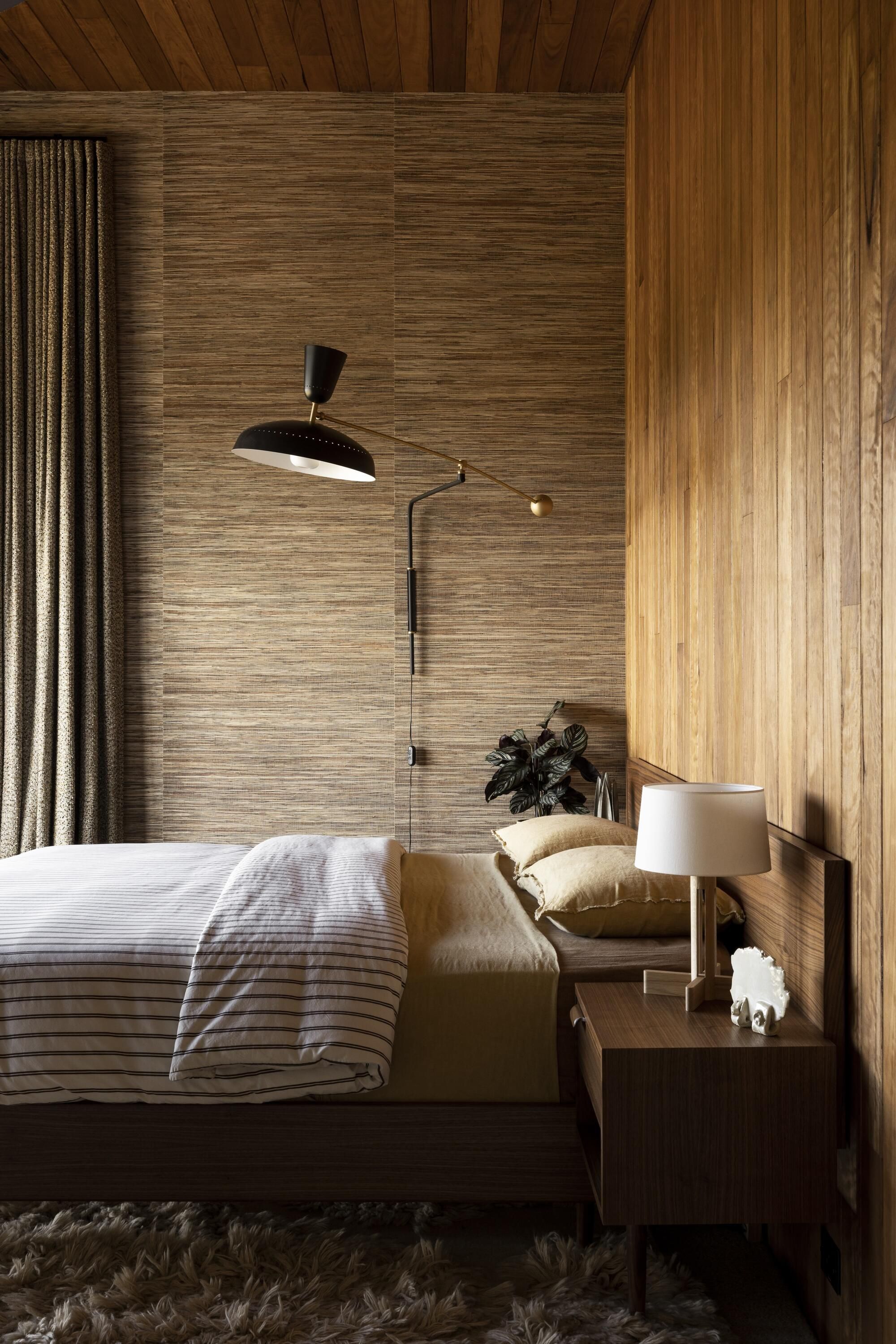 mid-century modern bedroom with textured wallcovering, timber panel ceiling and wall, striped bed linen on caramel colored sheets, drapes, fluffy rug, and timber side tables with lamp