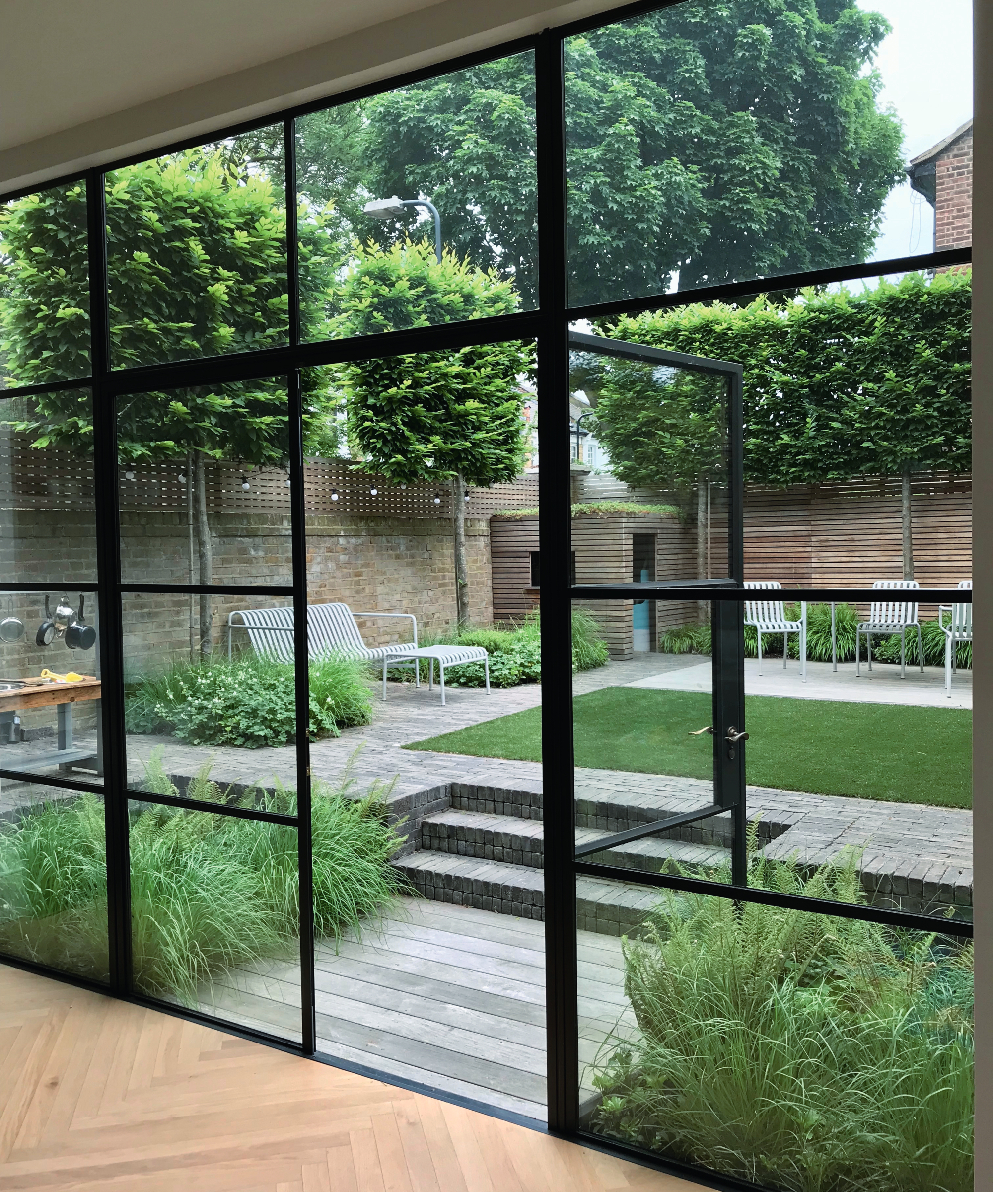 crittal style patio doors and windows leading to small garden with decked area leading to steps, lawn and paved areas