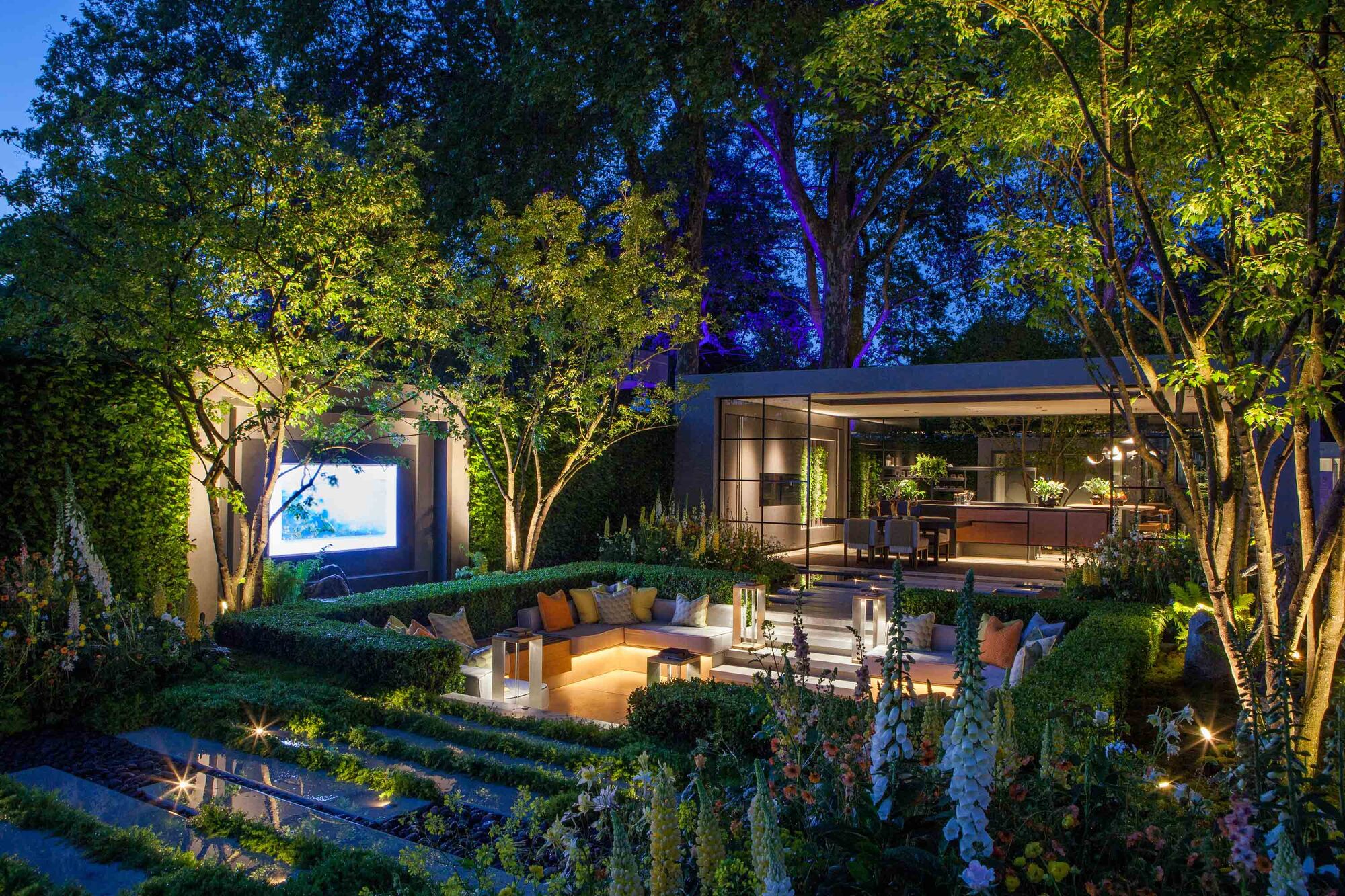 A modern garden at dusk with a conversation pit with lighting integrated into the seating, up-lit trees, and a view into a garden room with large windows