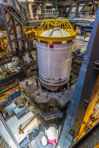 a tall silver cylinder stands upright in a very tall hangar on top of a rocket booster