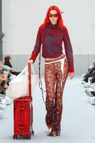 A female model with red hair wearing a blue and red tinted denim jacket and floral lace pants rolling her red luggage case down a runway.