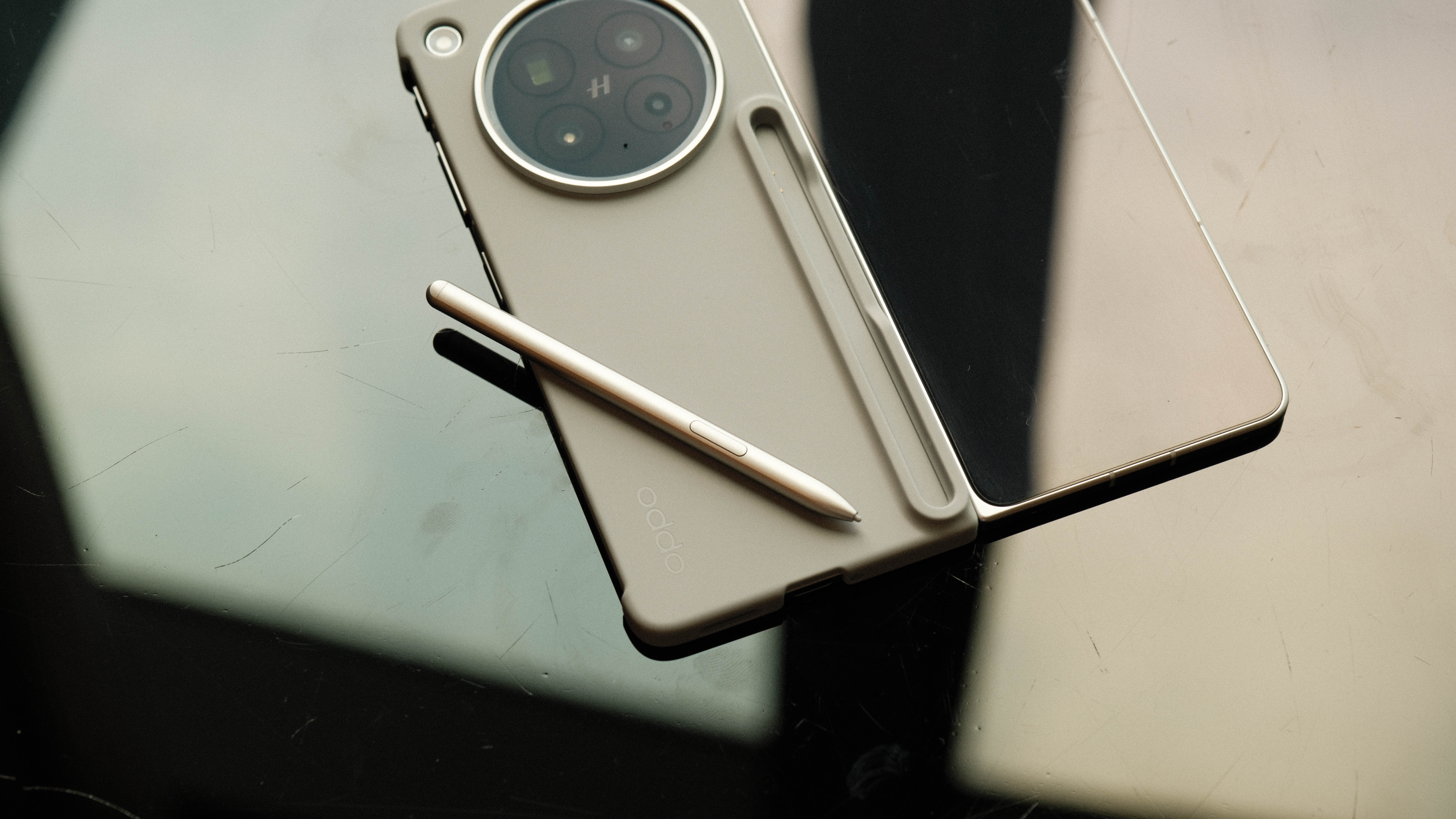 The Oppo Find N6 lying unfolded on a dark reflective surface. The matching silver stylus is resting on top of the tan protective case, next to its dedicated storage slot.