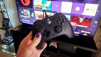 A hand with purple-painted nails holds an Xbox controller in front of a TV displaying a gaming interface with apps like Netflix and YouTube. The setting conveys a cozy gaming atmosphere.