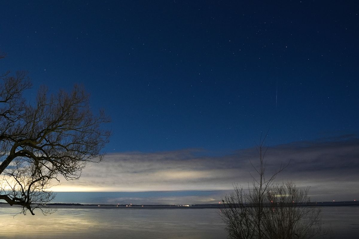 A meteor streaks across the night sky over Lake Simcoe, low cloud blocks the bright moonlight and a tree is in the foreground