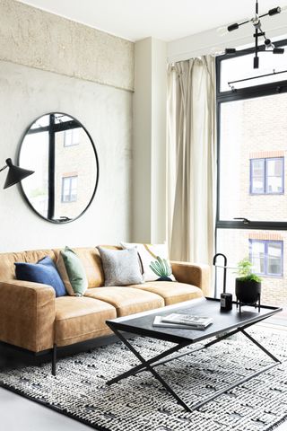 Mid-century modern lounge with leather sofa, neutral drapes, and large round wall mirror.