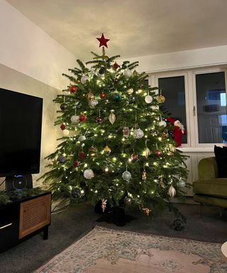 A large green natural Christmas tree in a neutral living room with autumnal decor and baubles, and a red star on top