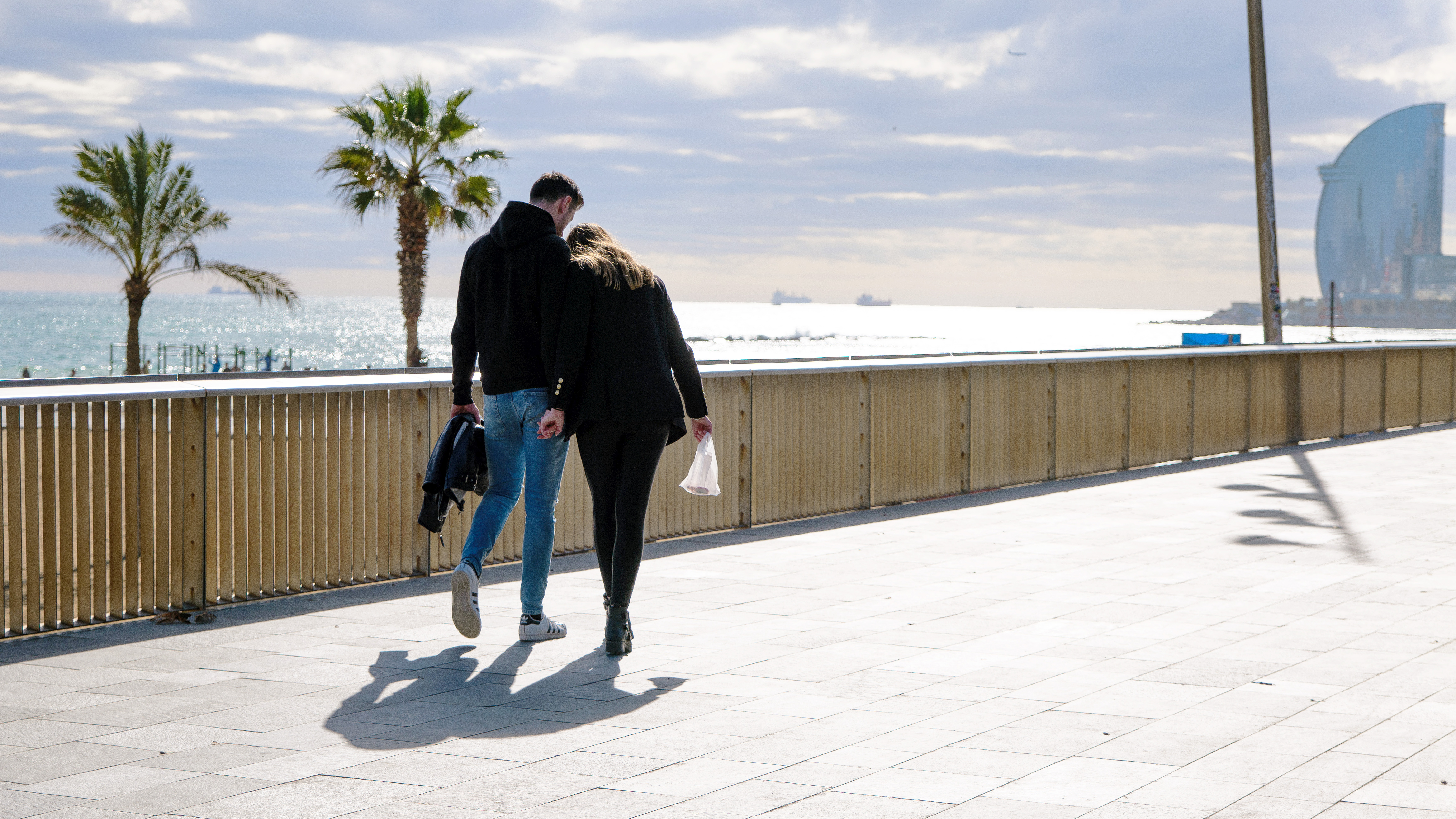 A couple walks on the beach in Barcelona with palm tress and ocean in the distance