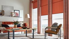 Colorful living room with large red curtains and blinds, red sofa, wooden coofee table and armchair, white rug, and misc art on a floating white shelf.