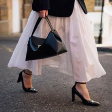 There is a zoomed in photo of a woman is wearing a white skirt and holding a black handbag and walking black heels.