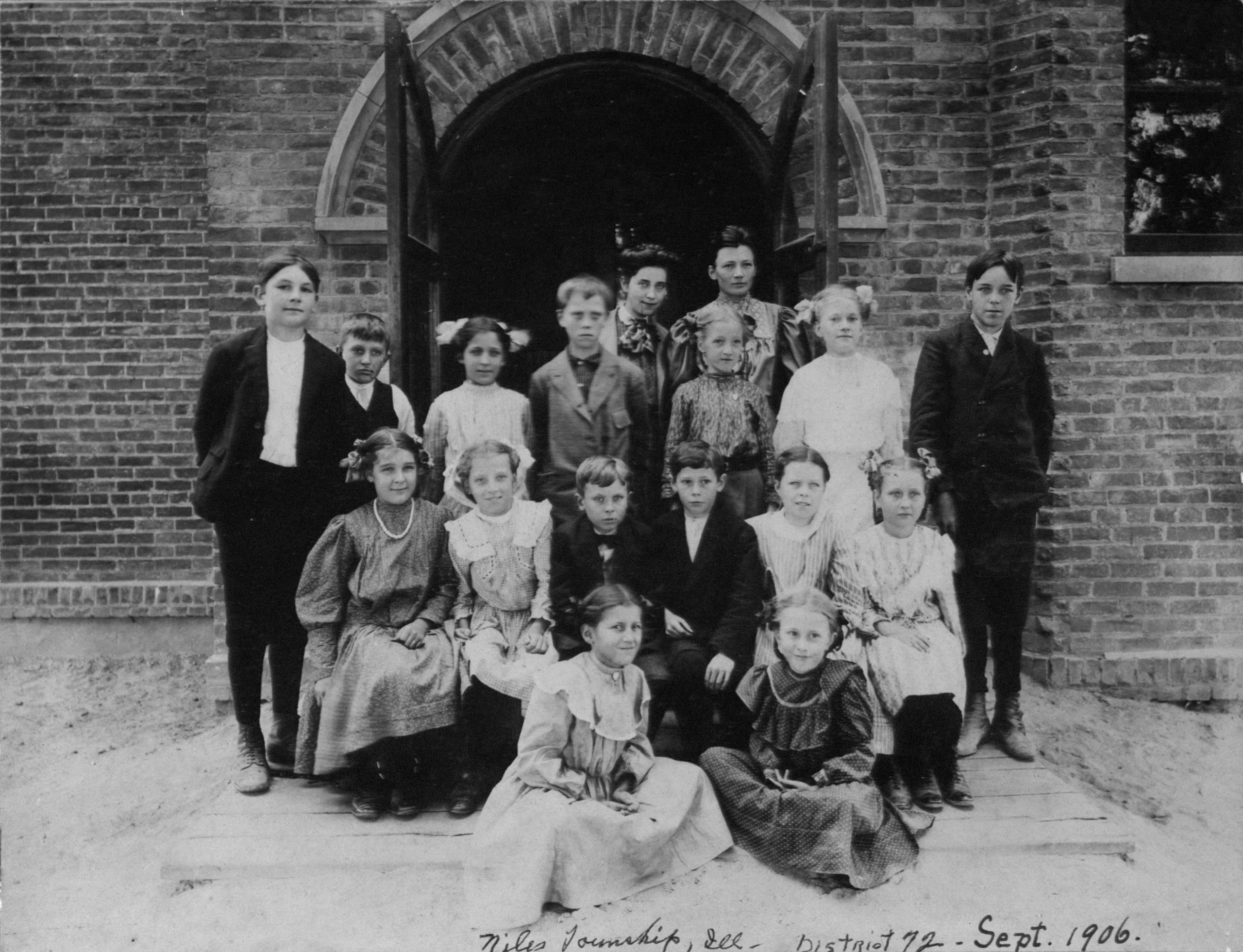 Around 20 children in high-collared Edwardian clothes pose in rows on the steps of an arched brick school doorway, with two women teachers standing at the back