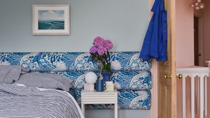 light blue bedroom with blue and white upholstered headboard, blue striped sheets, an artwork hung over the bed, a yellow door with a blue jacket hung on the back, and pink hallway walls 