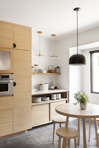 Scandi style white and blond wood kitchen with floor to ceiling cabinetry and open shelving, pendant lights, round dining table, stools