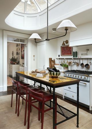 a skinny black metal and tile island sits in the middle of a small kitchen. Above the island are a pendant lighting with lamp shades. There is also a gas stove opposite the island with hanging pots and pans.