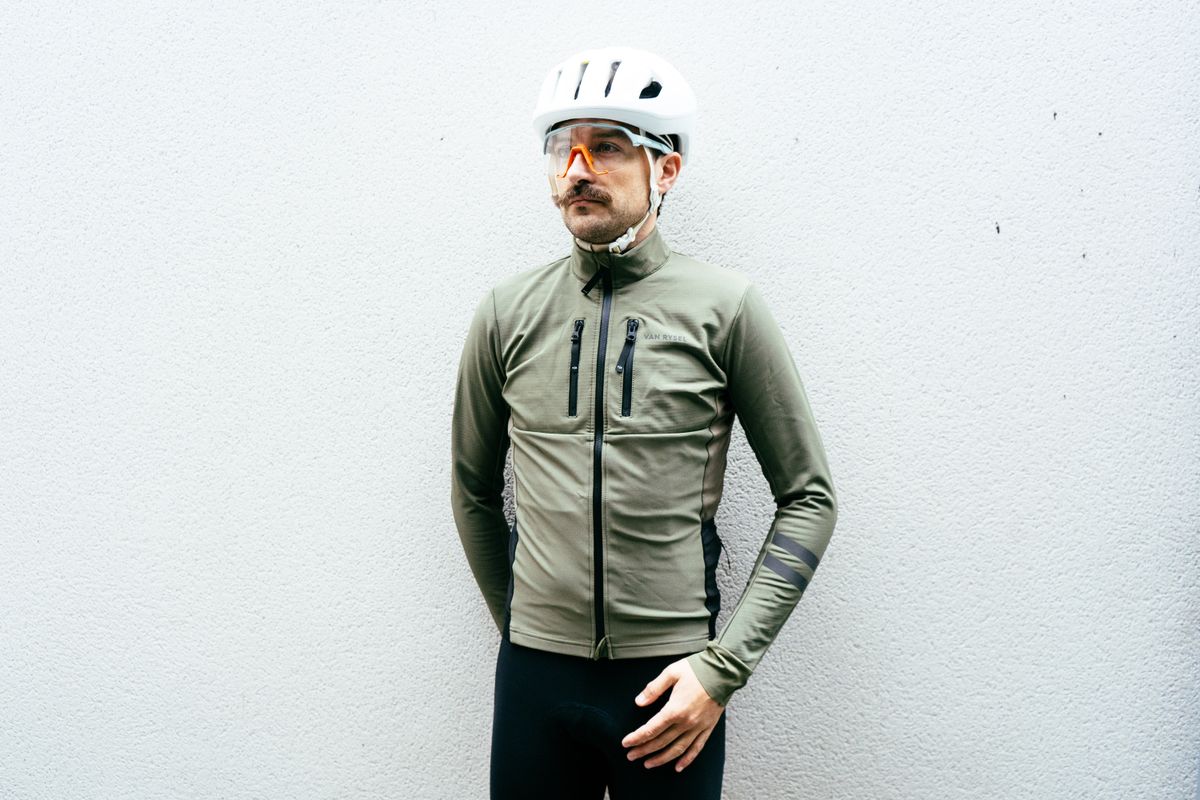 A white man in an olive green winter cycling jacket stands against a grey wall