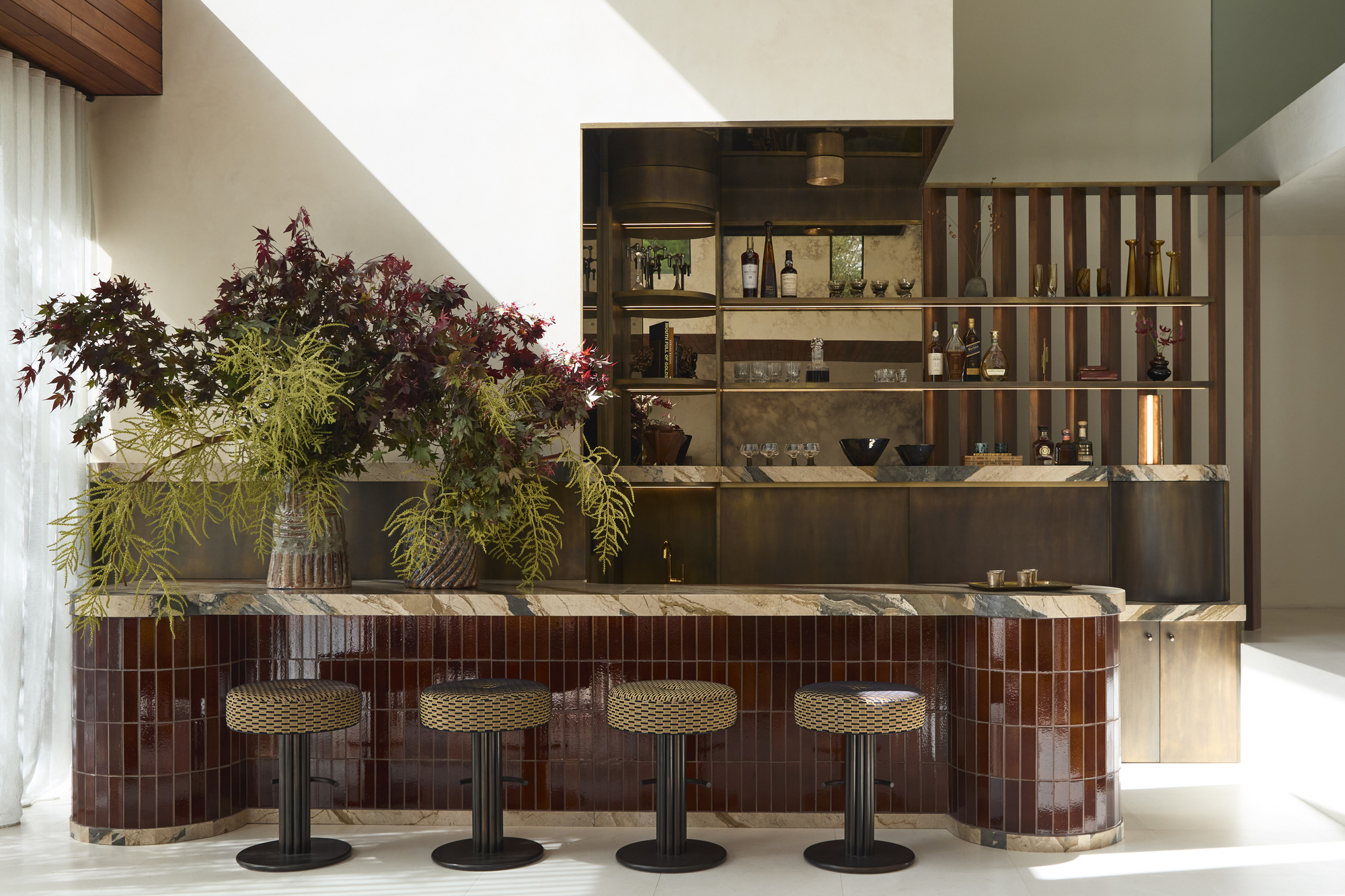 home bar with tiled island with marble top, four bar stools, a large floral display, and timber screen behind with built-in shelving, floating shelves with a mirrored back and white walls