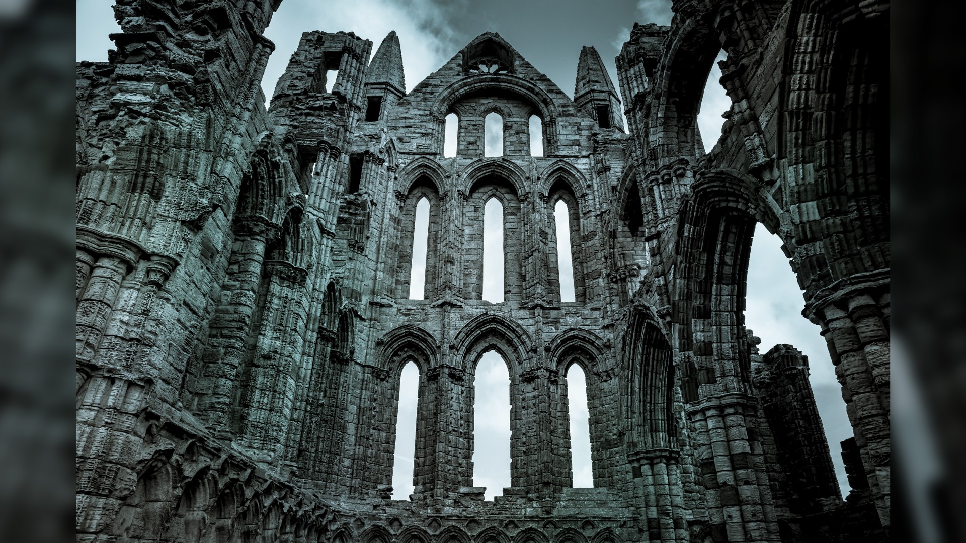 The dramatic ruins of the 7th century Whitby Abbey on the coast of North Yorkshire, England. The imposing building is made out of gray stone bricks, with numerous archways. We are facing a 3 by 3 layout of tall, thin window arches that get shorter as it goes up.