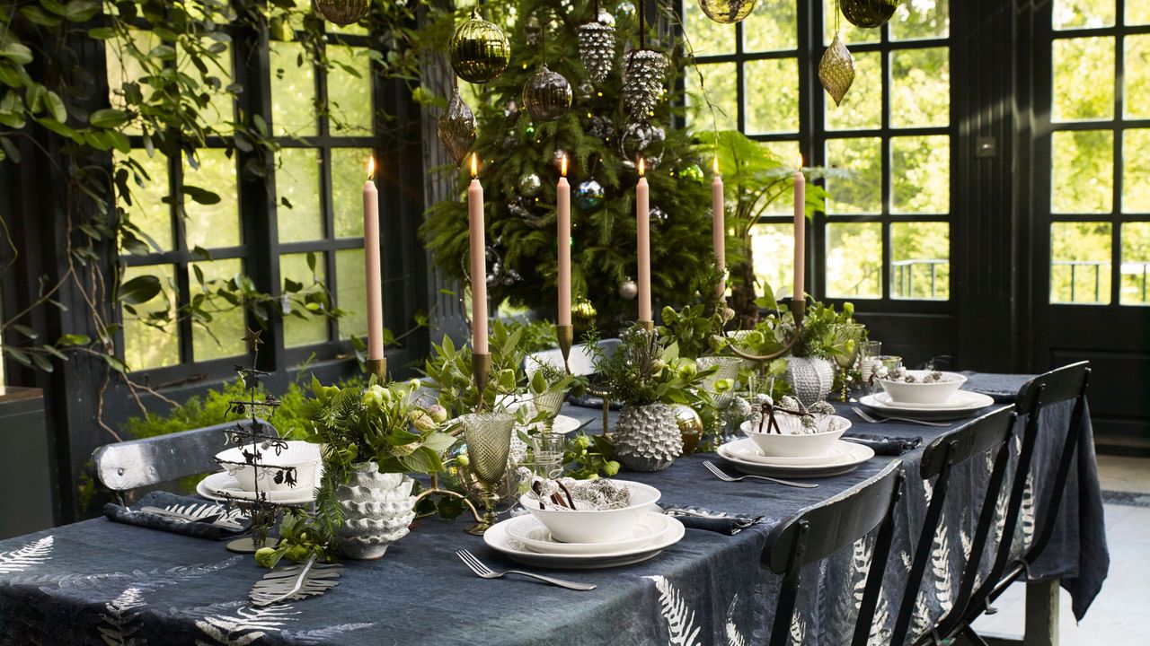 table setting in greenhouse with black tablecloth, white dinnerware, tall taper candles, and fresh green foliage