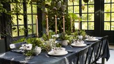table setting in greenhouse with black tablecloth, white dinnerware, tall taper candles, and fresh green foliage