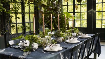 table setting in greenhouse with black tablecloth, white dinnerware, tall taper candles, and fresh green foliage