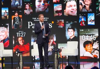 Prince Harry standing in front of a screen with pictures of people's phones and talking into a microphone