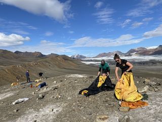 in the foreground on a hill are two people unfurling canopies and equipment. far in the back are other people with their own gear. mountains and blue skies with clouds are in the background, along with a little snow