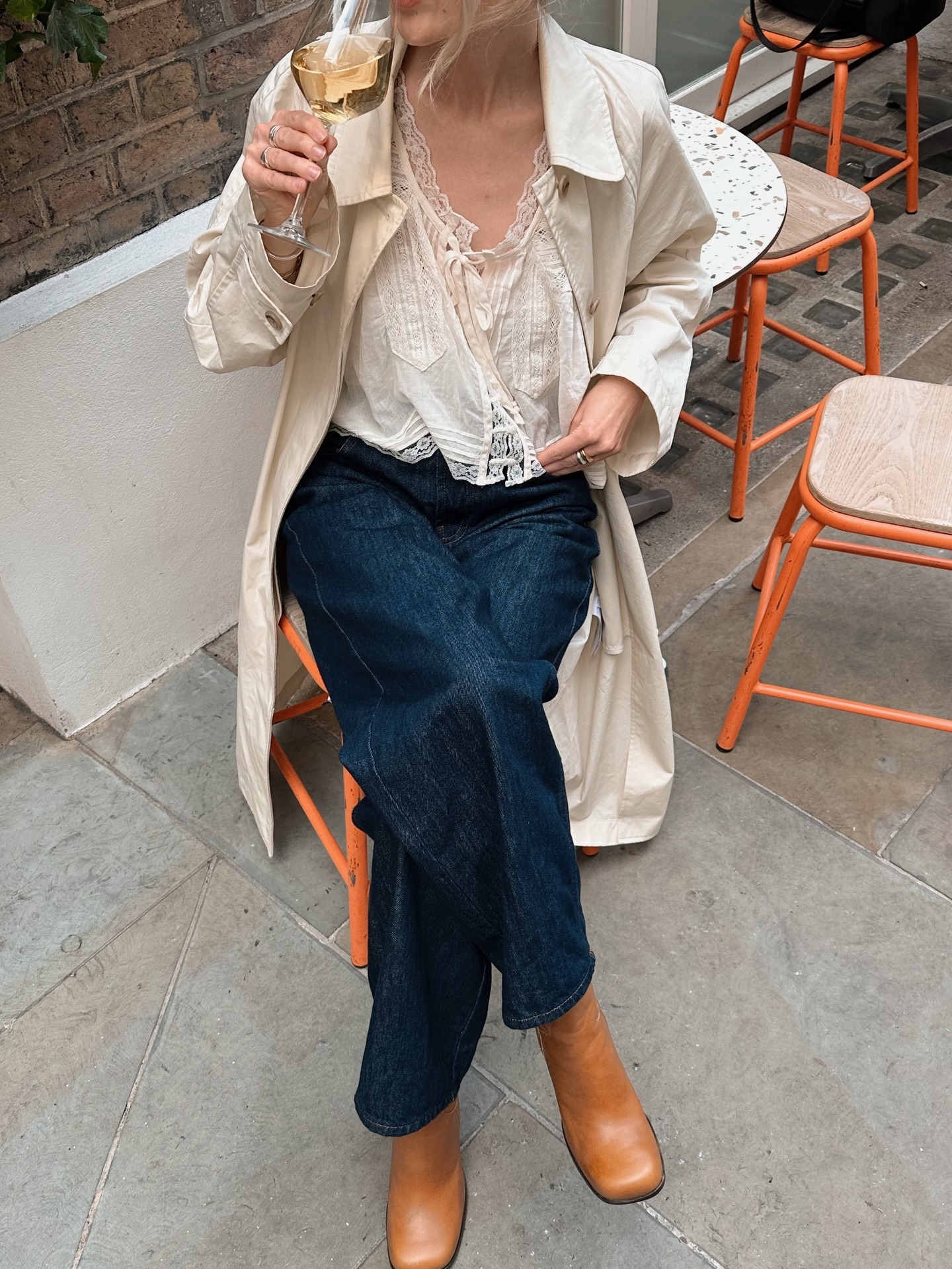 Fashion person Chloe Kath Butler wears a transitional winter to spring outfits 2026. She wears a embroidered blouse, blue jeans and brown boots. She has a cream trench over her shoulders. She is sat down, looking to the side drinking prosecco in a recent image from her Instagram.