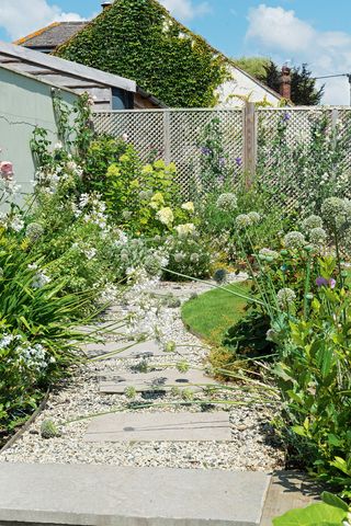 ideas for awkward shaped gardens: A garden path of pavers and gravel, with beds planted with alliums and flowering plants, and flowering plants on a trellis fence.