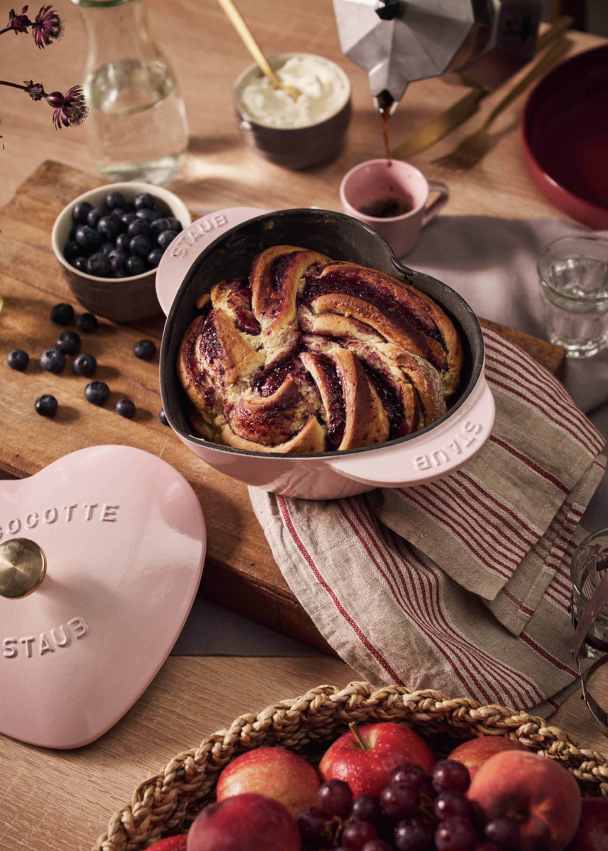 A dusty pink heart-shaped cast iron pot with a baked berry bread on a washcloth on a chopping board near bowls of berries and fruit