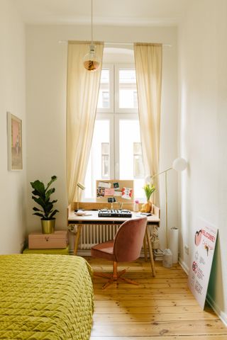 a small office at the end of a light airy bedroom with a wooden floor – a pink desk chair, and a green/yellow bedspread, with a high window