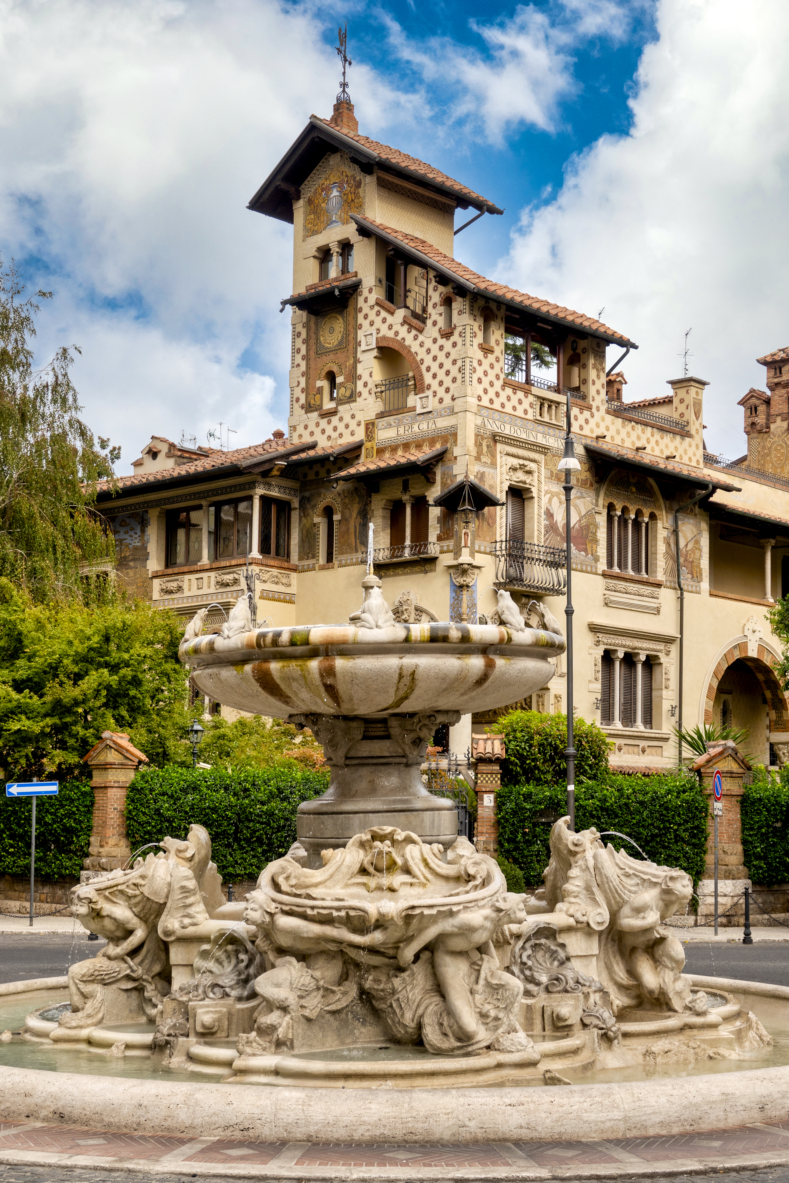 An esoteric fountain depicting multiple human silhouettes along with sea life and shell-shaped motifs in a leafy residential area with beautifully ornate architecture.
