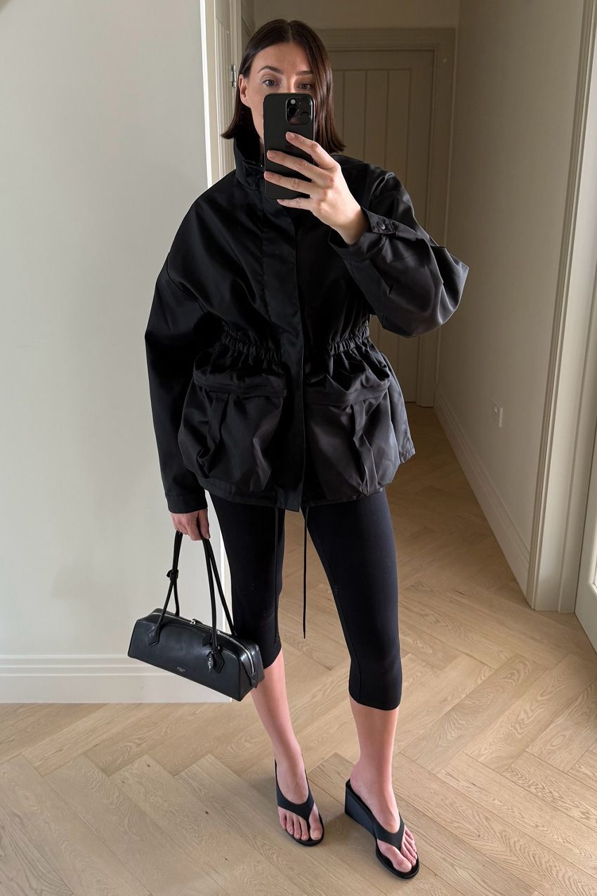 A windbreaker and plain leggings outfit idea is shown in a photo of a woman taking a mirror selfie in a minimal hallway of their home wearing a large black parka with black cropped leggings, black thong heeled sandals, and a Le Tekel Alaia bag