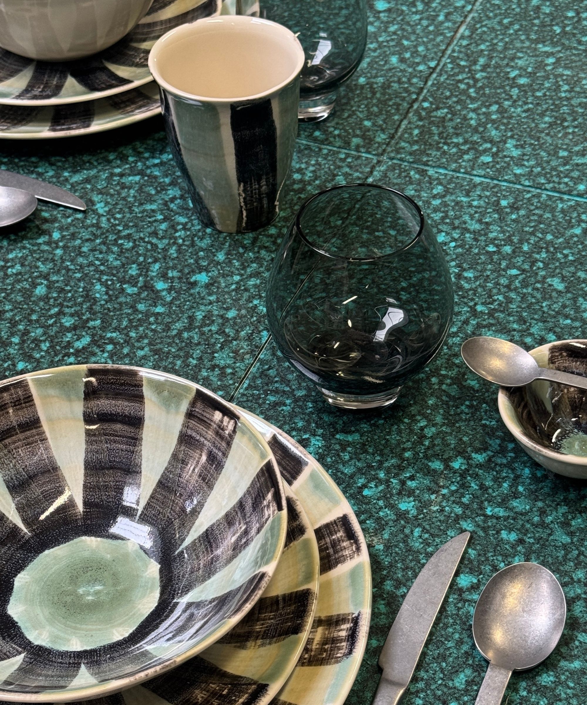 handpainted striped dinnerware set with plates and bowls, silver cutlery, green glass, ceramic cup, on green speckled ceramic tabletop