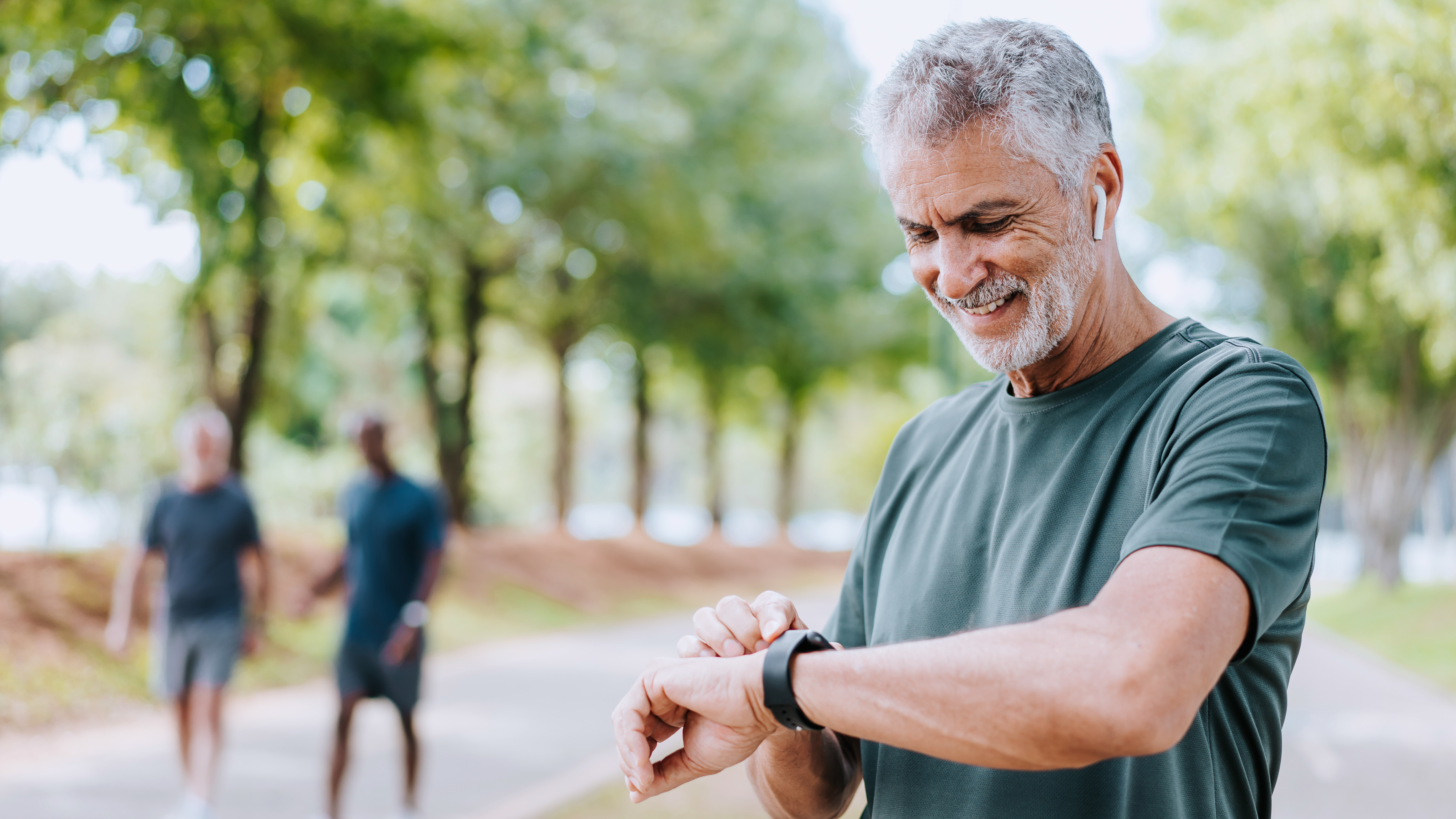 A man stands in a park smiling and looking at his smartwatch.