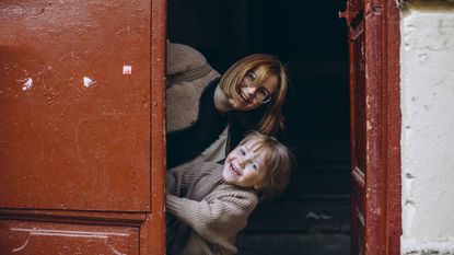A mother and young son peek playfully from behind a large, red door.