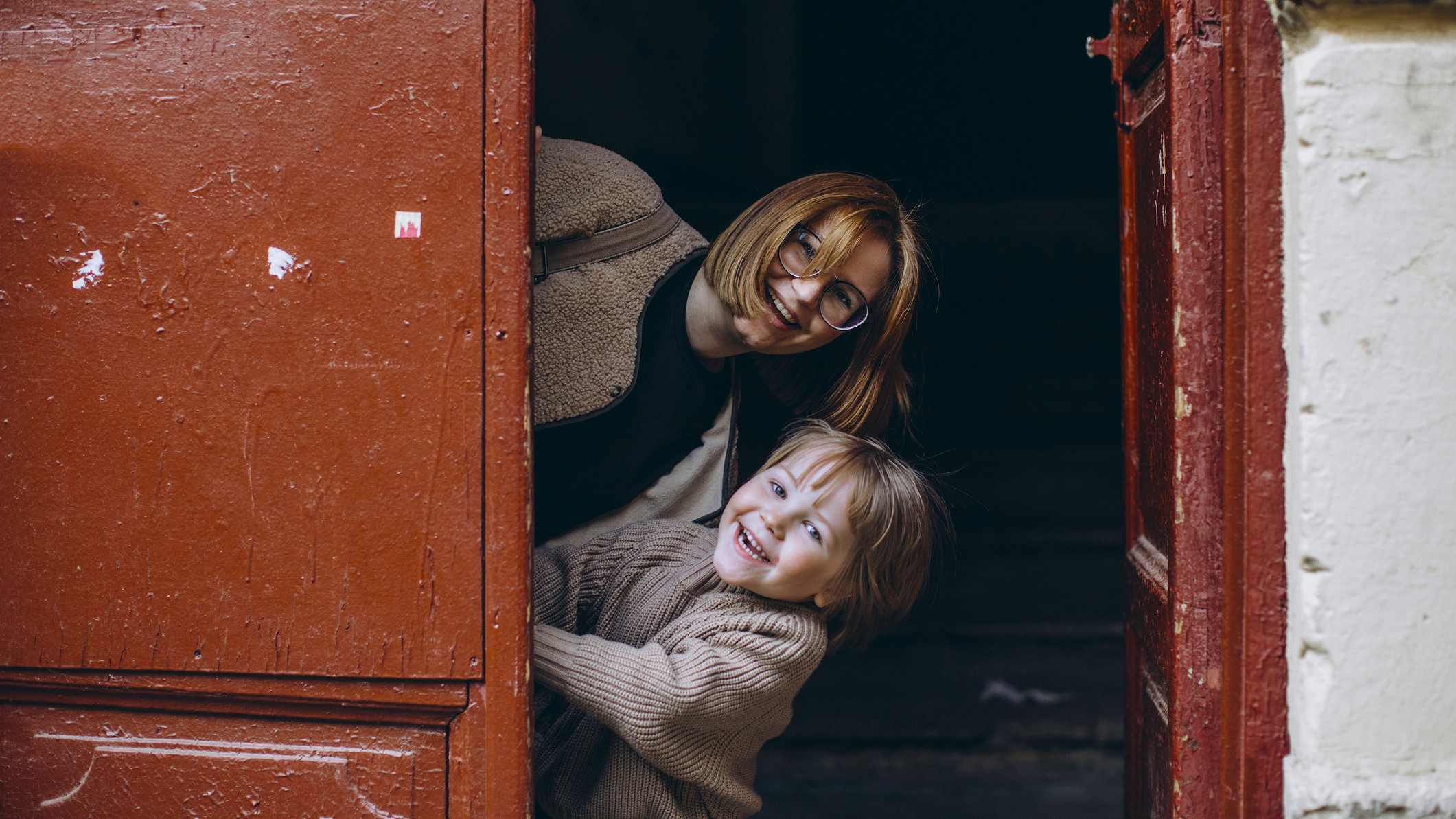 A mother and young son peek playfully from behind a large, red door.