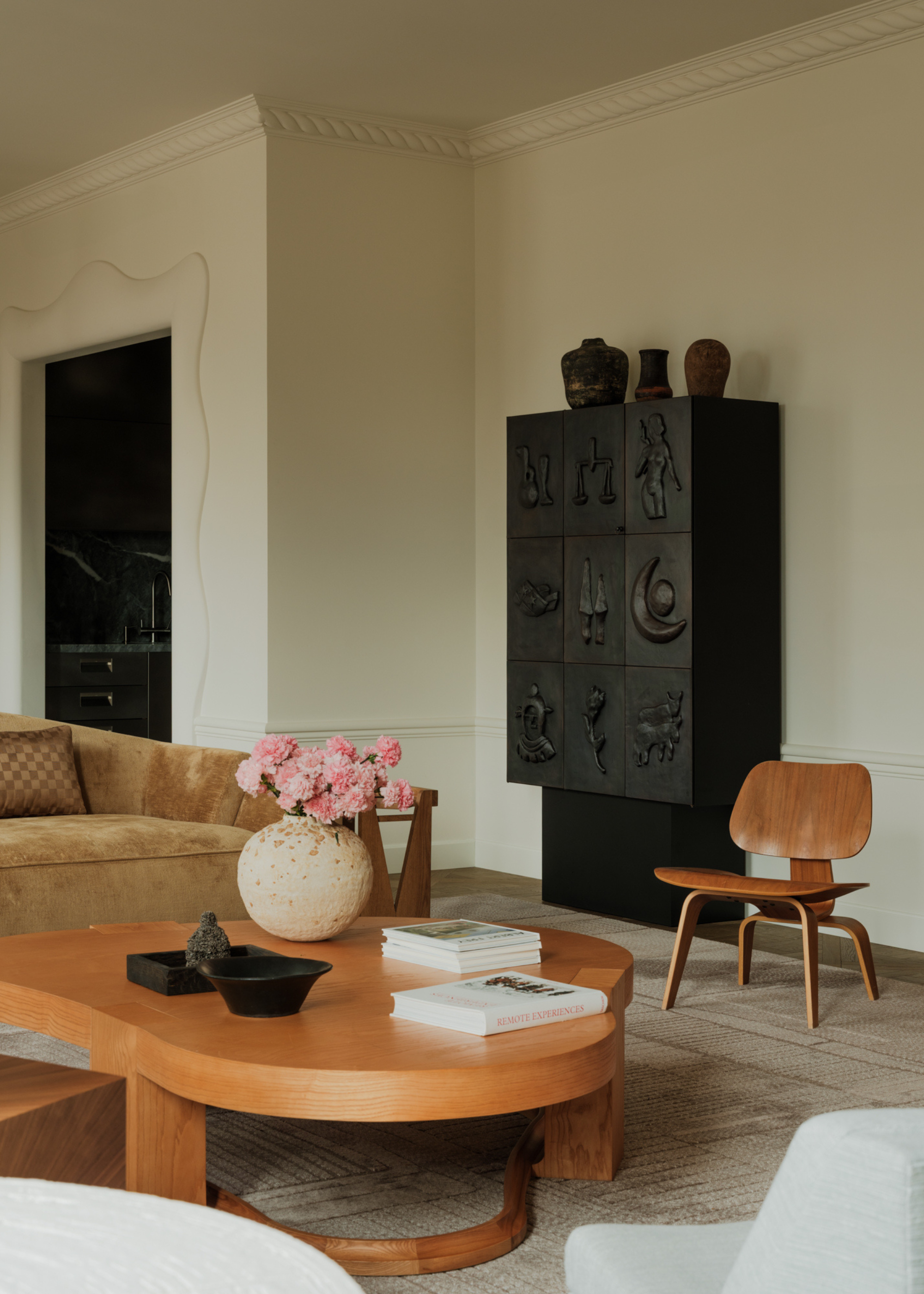 A white living room with a black debossed cabinet by a wood chair, a velvet couch, a low curved coffee table, and a vase of pink flowers